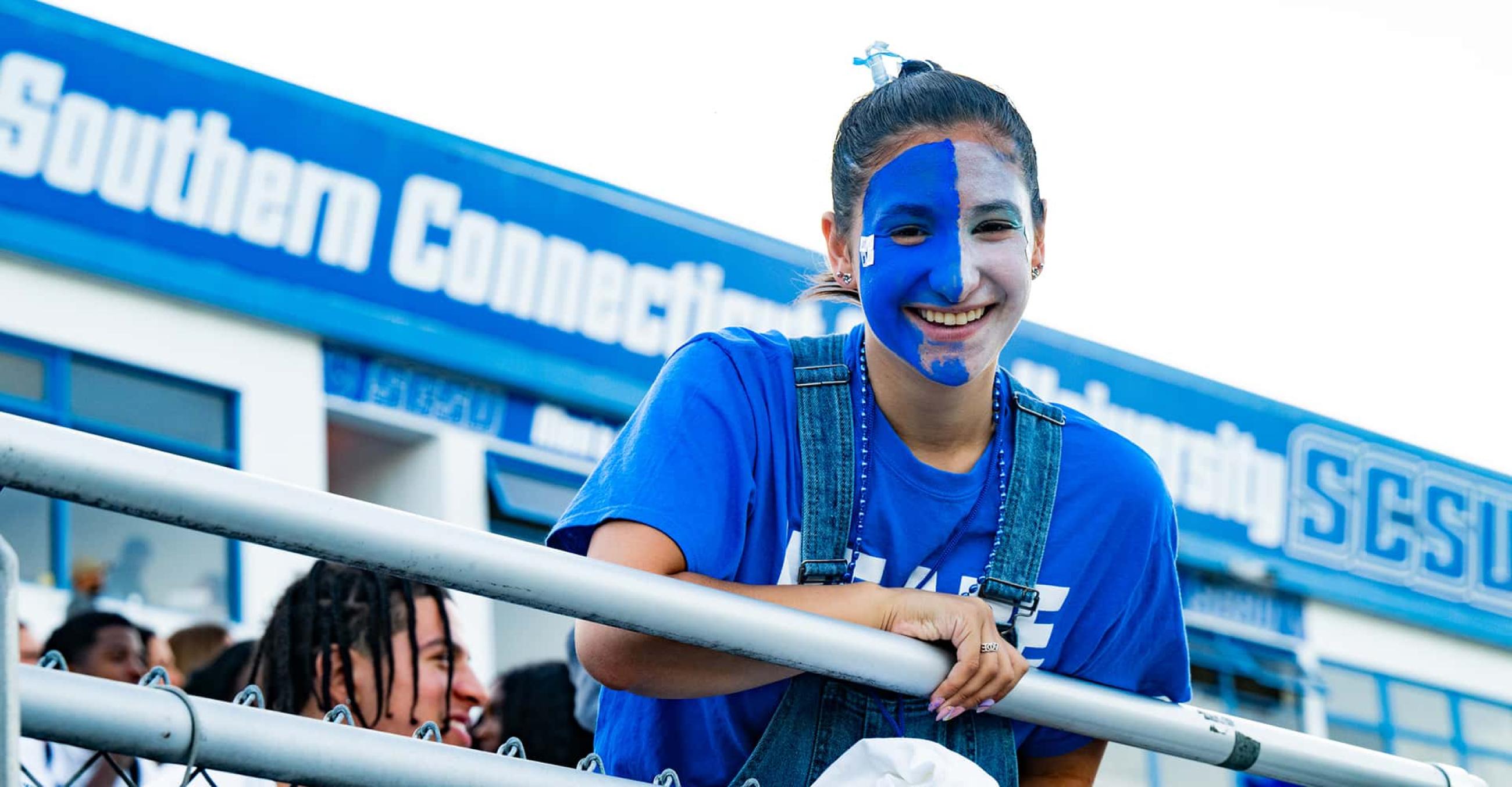 a student with her face painted blue and white smiles down at the camera from the bleachers at a football game
