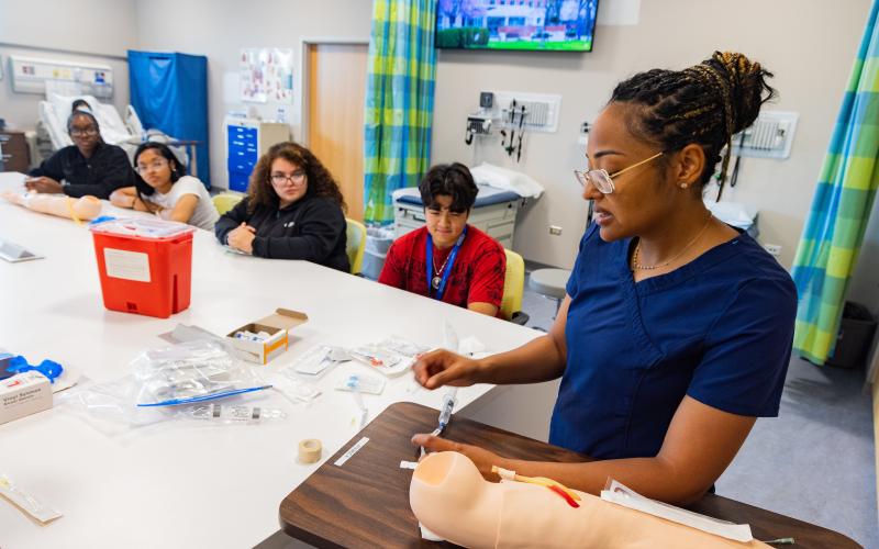 A nursing instructor in navy blue scrubs demonstrates how to insert an IV using a medical training arm, while a group of attentive high school students watch during a hands-on learning session in a clinical simulation lab.