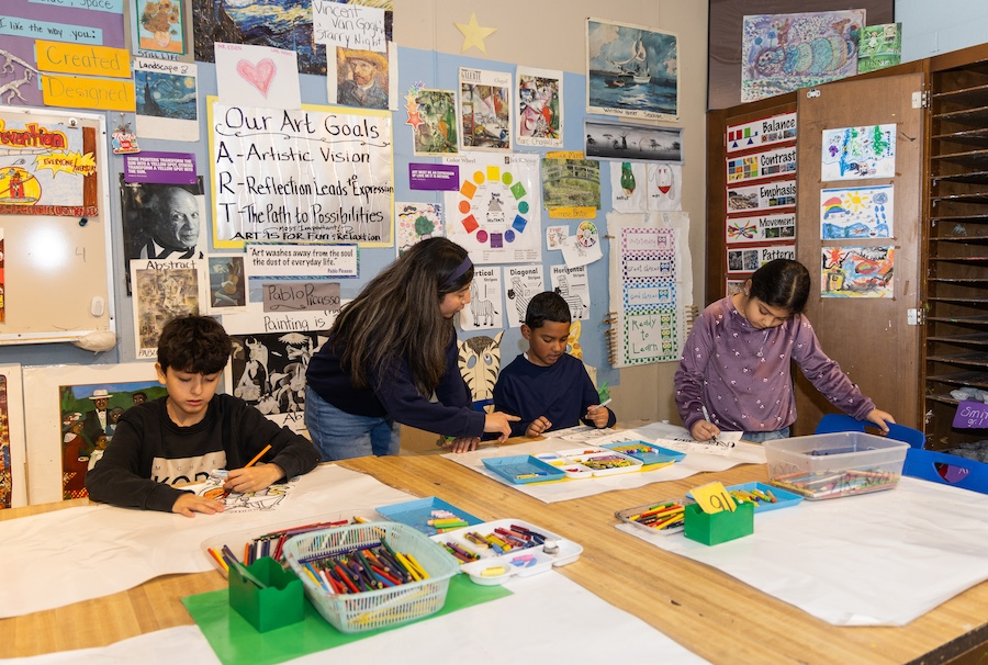 A student teacher working amongst three elementary students at a table, helping them with work.